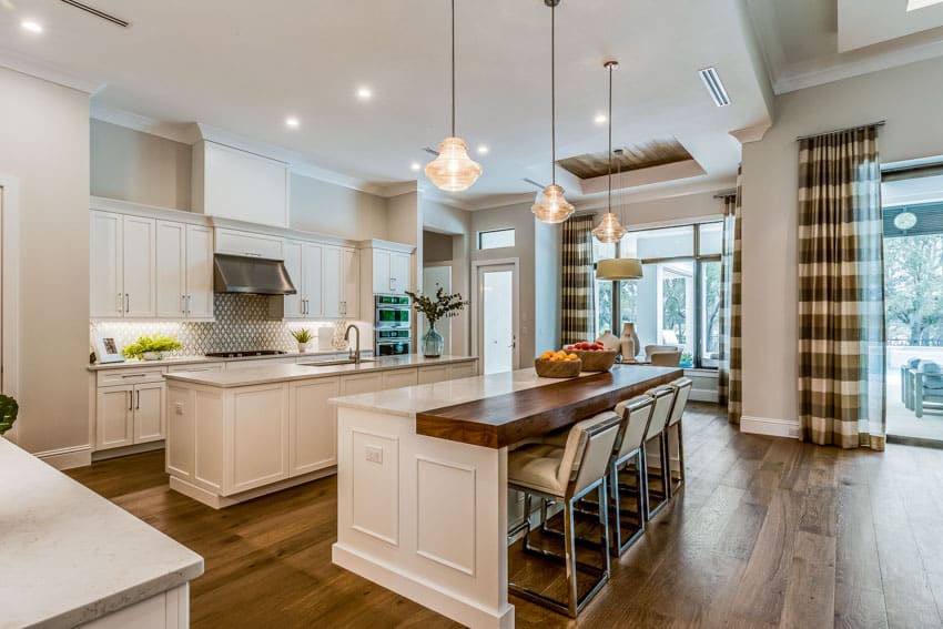 Kitchen with island, built in cutting board, chairs, white cabinets, backsplash, range hood, pendant lights, glass windows, and curtain