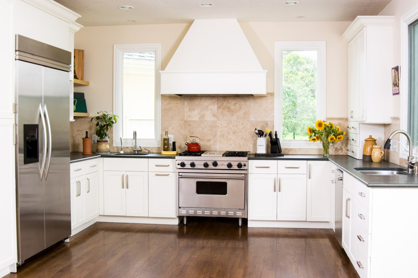 Kitchen with limestone tile backsplash, wood floor, white cabinets, range hood, and windows