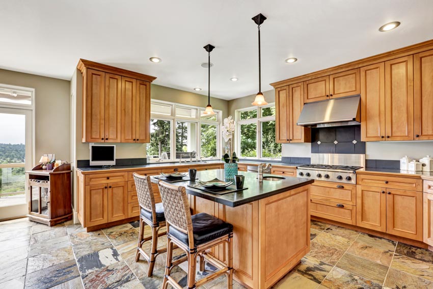 Kitchen with stone tile floors, peninsula, chairs, hanging lights, red oak cabinets, countertop, range hood, and windows
