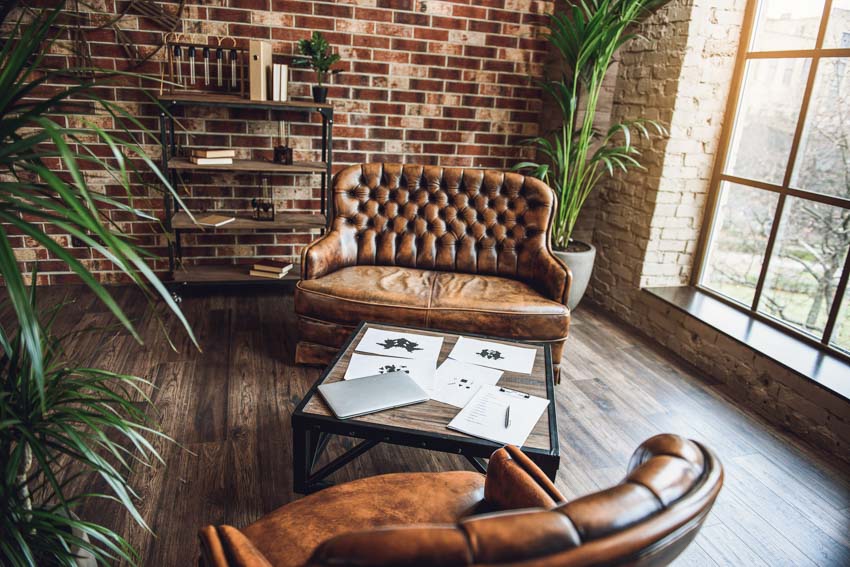 Living room with brown leather sofa, chairs, brick wall, shelves, wood floor, indoor plants, and windows