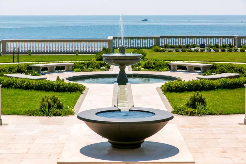 Luxurious landscaped area with benches, hedge plants, paved walkway, water features, and types of fountains