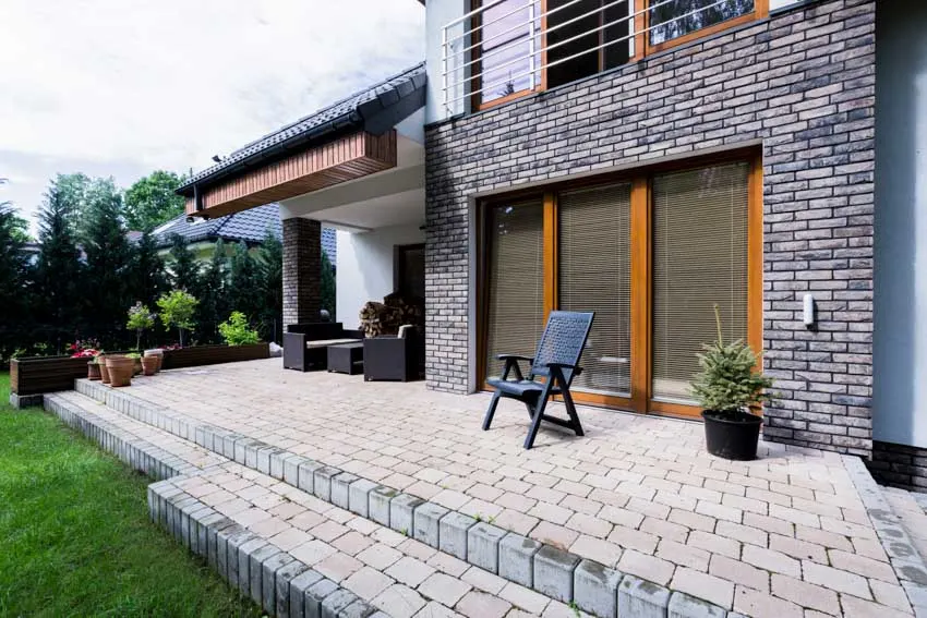 Paved patio surface with chair, brick house wall, windows, and potted plants