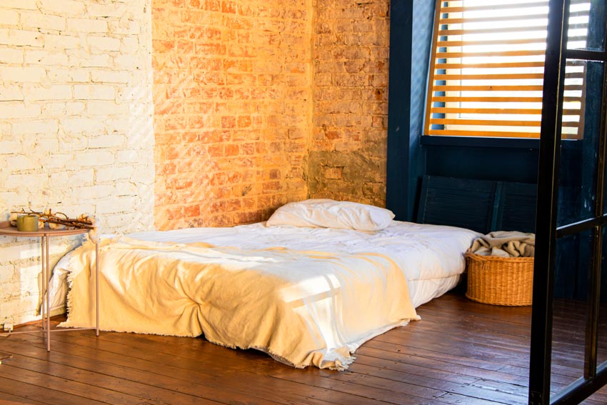 Simple bedroom with mattress, pillows, brick wall, wood floor, and window shutters