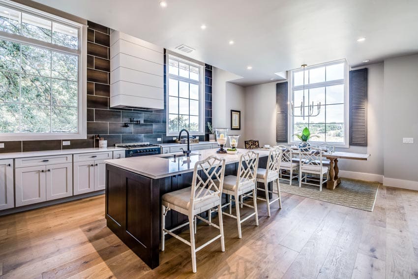 Spacious kitchen with subway design wood tile backsplash, island, chairs, countertops, range hood, wooden flooring, and windows