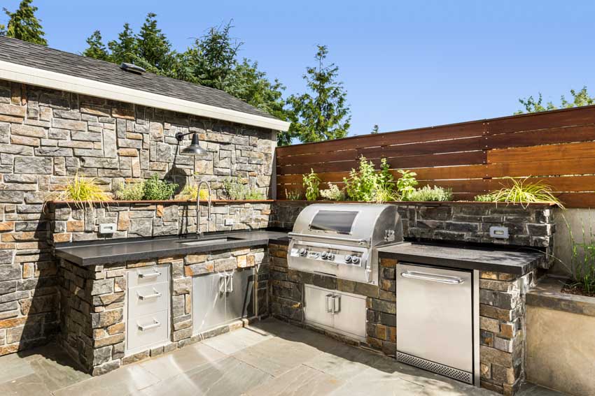 Stone outdoor kitchen with countertops, barbecue grill, backsplash, and wood fence