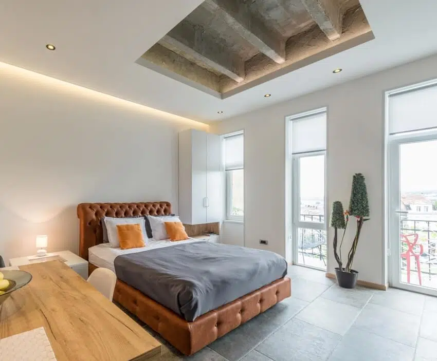 Bedroom iwith brown leather bed frame, concrete tile floors, potted plant near glass windows 