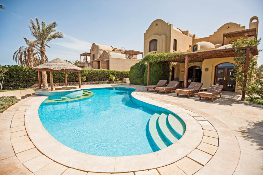 Beautiful villa with pergola, lounge chairs, pool, hedge plants, trees, and limestone pool deck