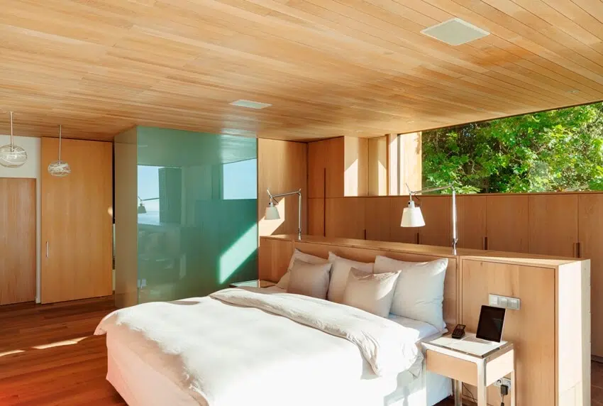 Bedroom with white cozy bed, window with a view of trees and wooden paneling ceiling 
