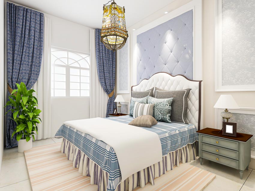 Bedroom with bed skirt, comforter, headboard, pillows, nightstand, lamp, indoor plant, window curtains, and rug