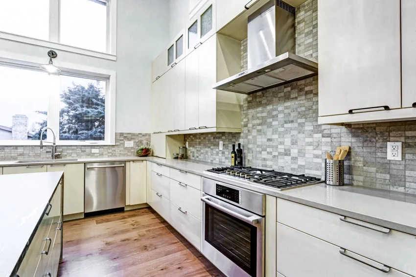 Kitchen with onyx mosaic tile backsplash, cabinets, range hood, countertop, windows, and stove