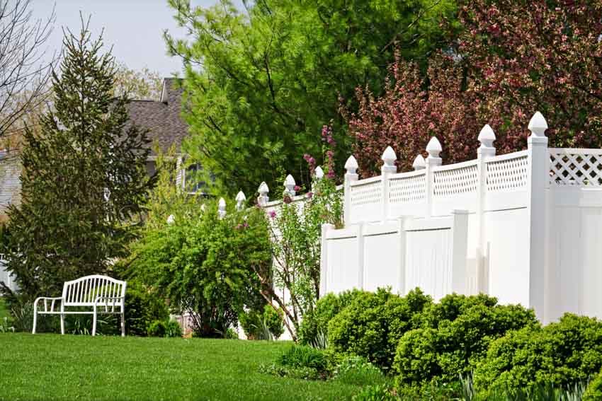 Outdoor area with white vinyl fence, bench, trees, and hedge plants