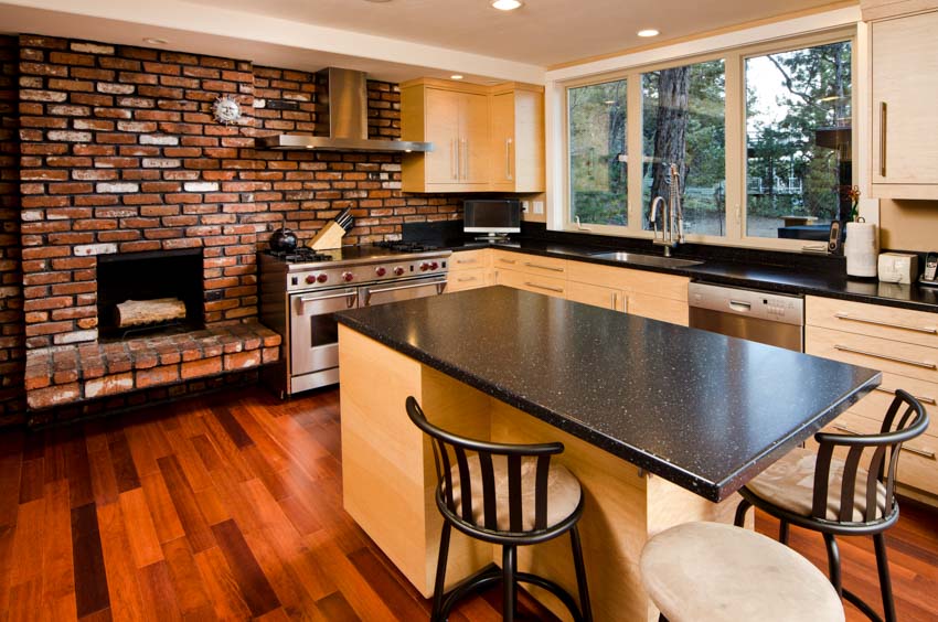 Rustic industrial kitchen with exposed brick backsplash, island, countertops, chairs, wood floors, cabinets, range hood, stove, and windows