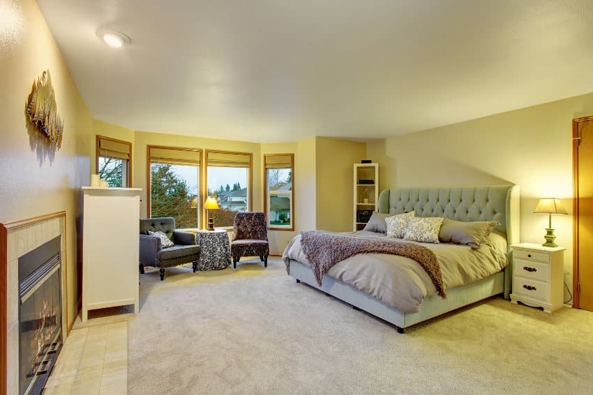 Spacious and comfortable bedroom with gray tufted bed, warm light, glass windows and gray carpet floors