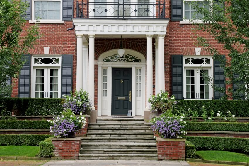 A big house made of bricks with nice landscape and a portico style entrance with dark colored door 