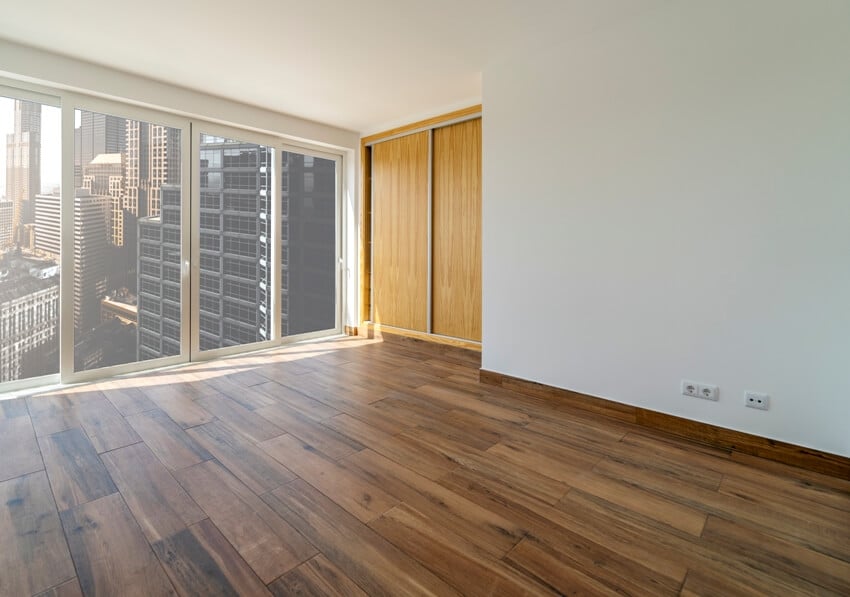 An empty room with dark floating laminate flooring and glass windows with a view of buildings