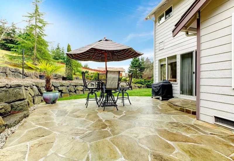 Flagstone flooring on cozy patio with iron table set and patio umbrella surrounded by green terrace landscaping