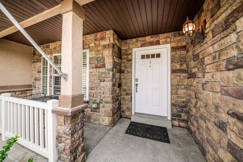 Front porch with composite outdoor ceiling panels, white door, stone wall cladding, and floor mat