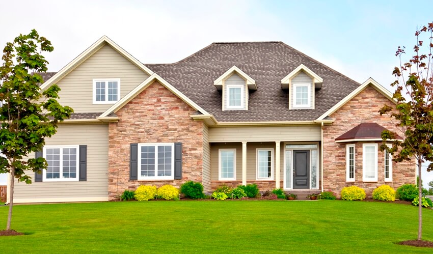 A gorgeous tan brick home with white windows, gray shutters and big front lawn with landscape and green grass