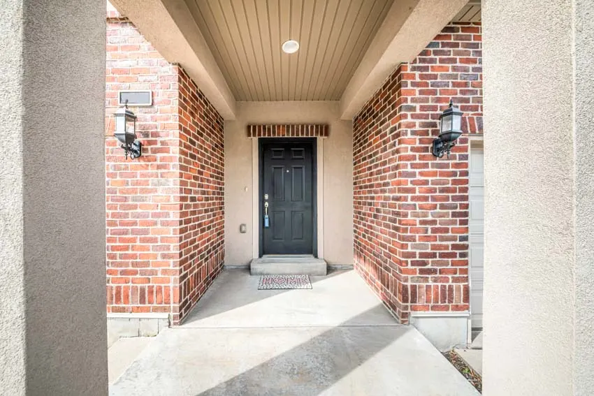 House entrance with black door, brick wall, outdoor ceiling panels, and wall sconces