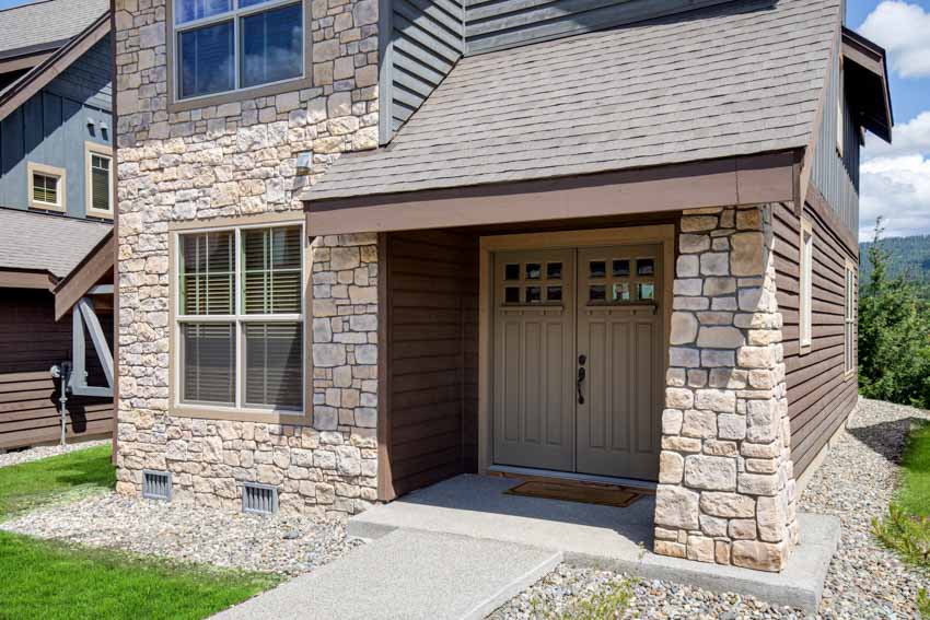 House exterior with double doors, castle stone siding, shingle roof, and lawn