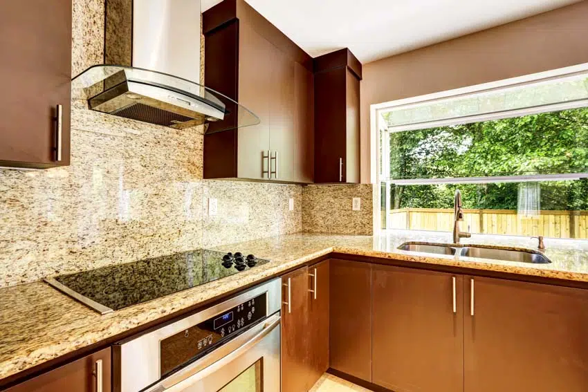 Kitchen with cream granite countertop, backsplash, stove, range hood, cabinets, sink, faucet, and window