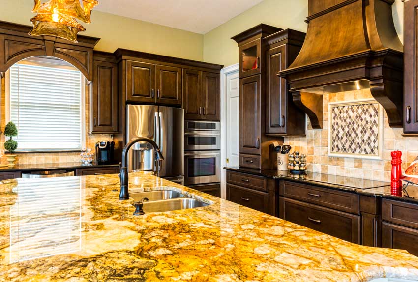 Kitchen with gold granite countertop, backsplash, cabinets, range hood, sink, faucet, and window