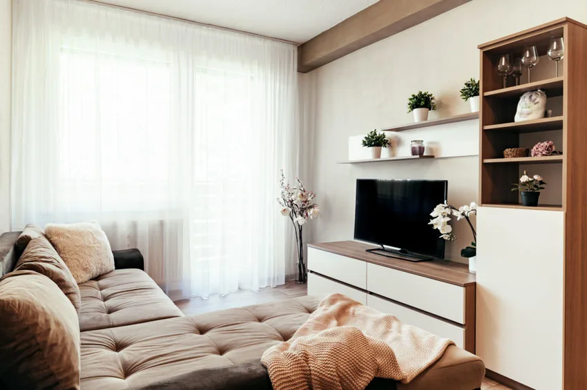 Living room with cream walls, brown furniture, curtains, floating shelves, television, and TV stand