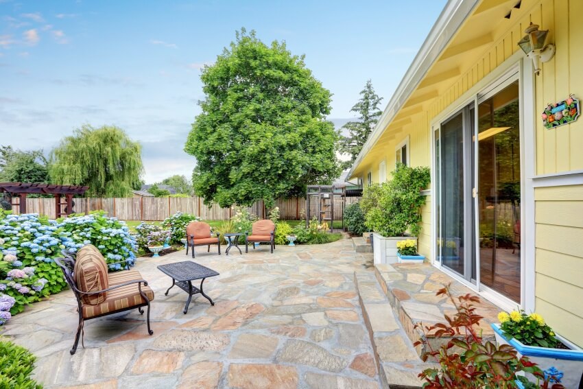 Perfectly designed patio area with flagstone floor in the backyard of a yellow house with relaxing comfortable outdoor furniture and blooming flowers