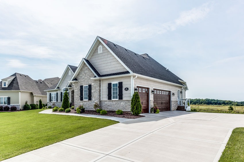 Residential house exterior with driveway, natural stone siding, pitched roof, hedge plants, windows, and lawn