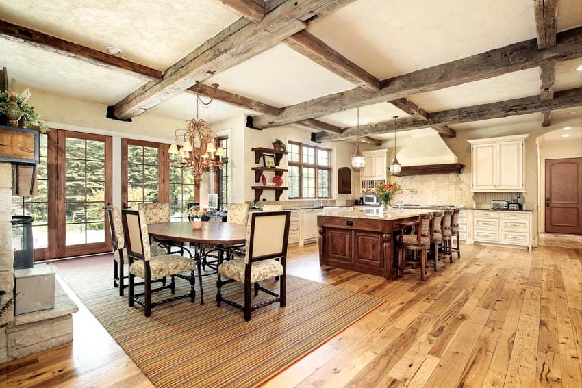 Rustic kitchen with chandelier, dining table, chairs, island, cabinets, range hood, ceiling beams, windows, and wood flooring