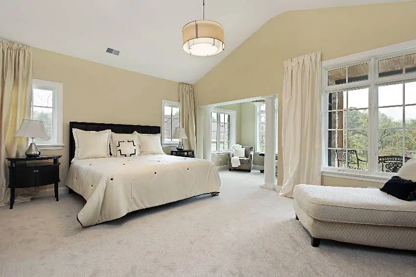 A spacious cream bedroom interior with natural lighting, carpet flooring, french windows and a gray couch near the windows