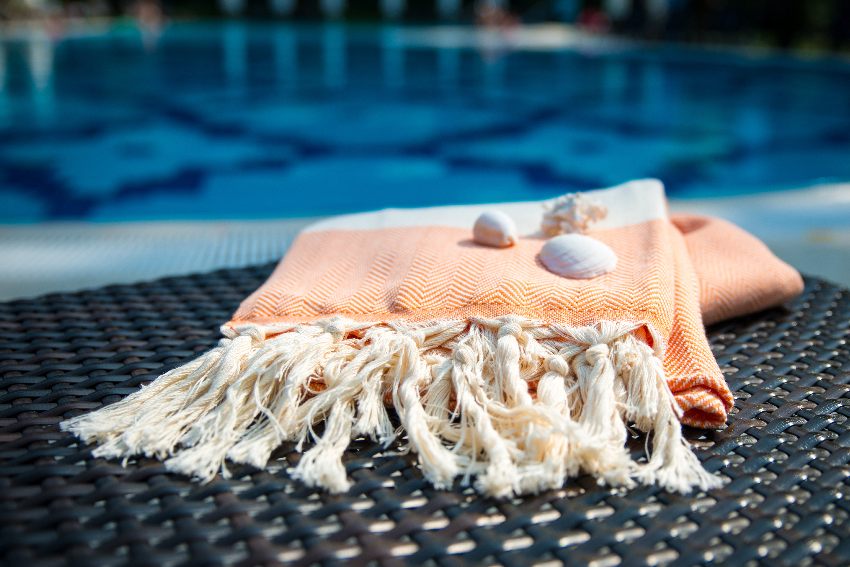 Summer accessories with white and orange Turkish towel and white seashells on rattan lounger with swimming pool at the back