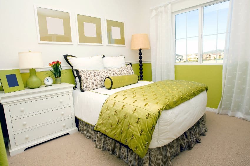 A white and lime green bedroom interior with bed, floor lamp and side drawer with lamp, alarm clock, photo frame and potted plant