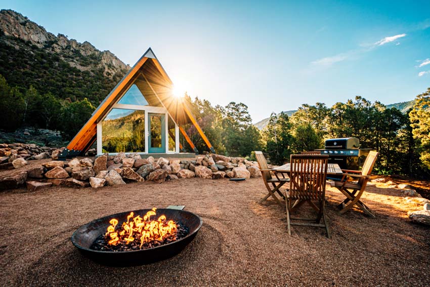 A frame tiny house with windows, fire pit, outdoor table, chairs, and landscaping rocks