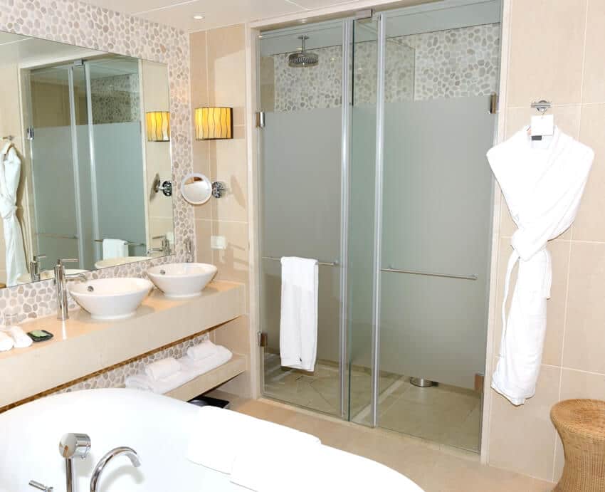 Bathroom interior with bathtub, two sink basins and partially frosted glass shower doors with bathrobe and towel beside it