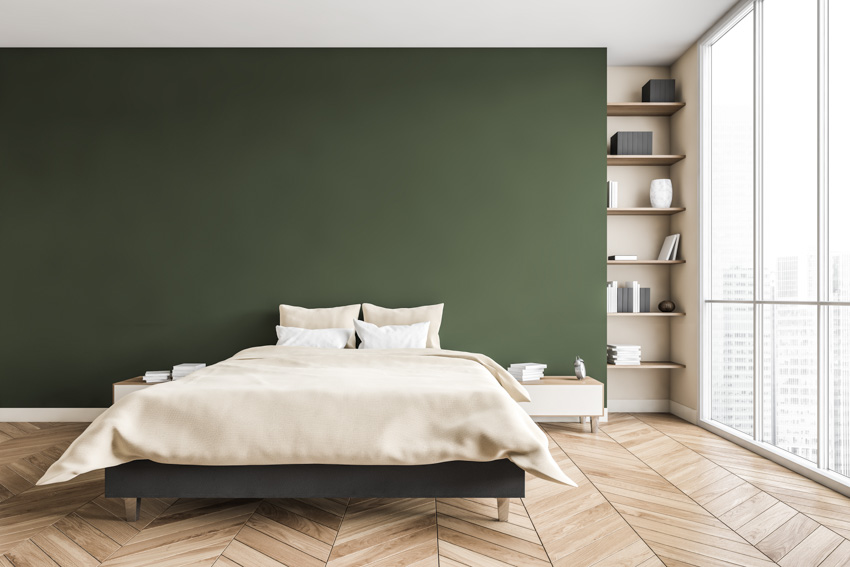 Bedroom with olive green wall, herringbone wood floor, bedding, pillows, shelves, and window
