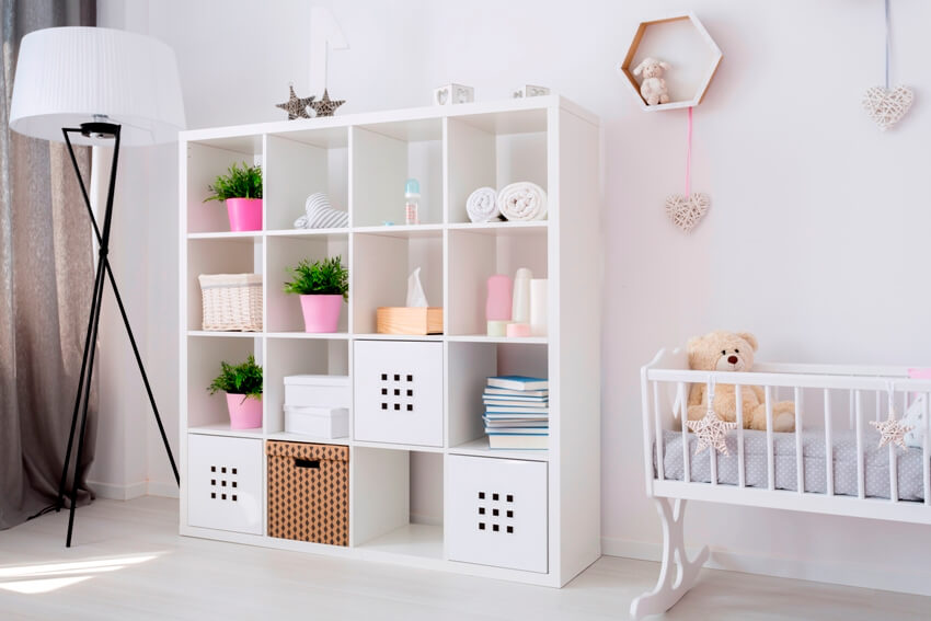A bright baby girl room features white cubby storage with baskets, baby cradle with a teddy bear and a lamp
