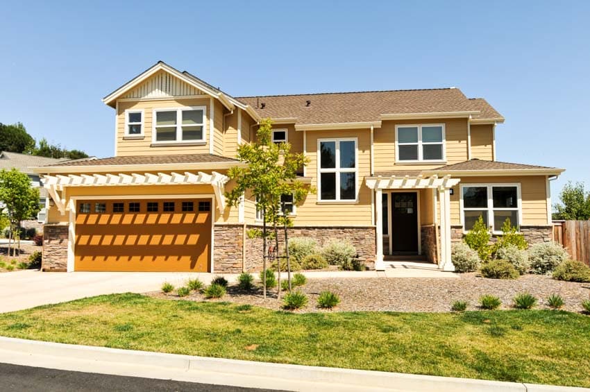 Brown and yellow house exterior with garage door, stone wall cladding, windows, front door, and lawn