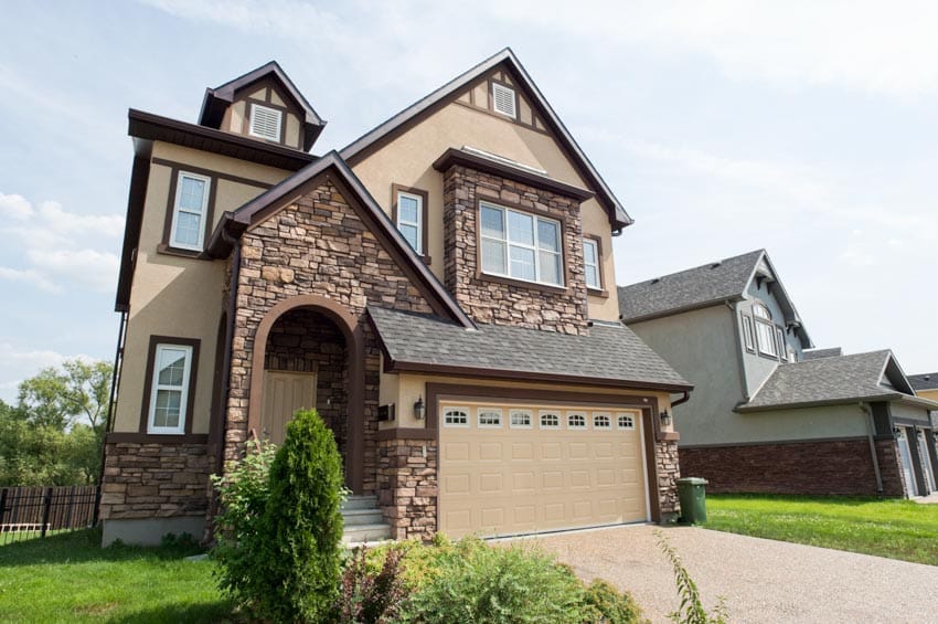 Brown house exterior with stone wall cladding, garage, driveway, plants, and windows