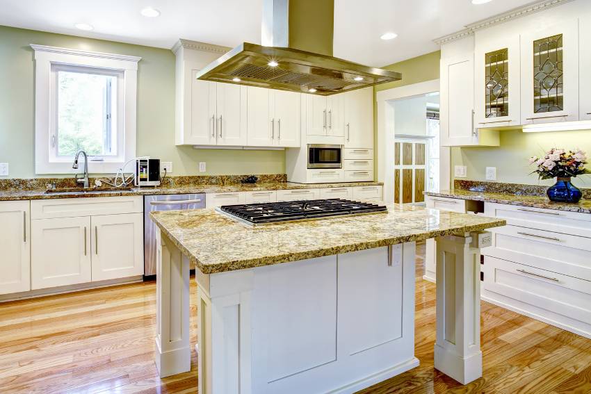 Kitchen interior with white cabinets, brown granite countertops, kitchen island with built in stove and steel hood