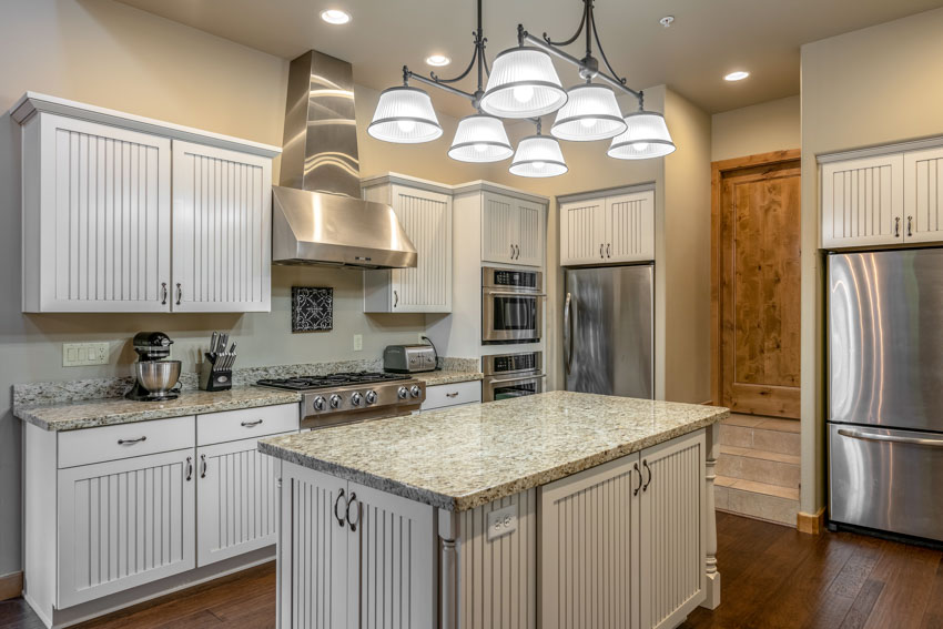 Kitchen with beaded recessed cabinets, countertops, island, pendant lights, backsplash, wood floor, and range hood