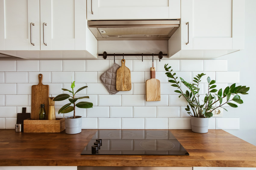Kitchen with ceramic tile backsplash, wood countertop, stove, range hood, cutting board, and cabinets
