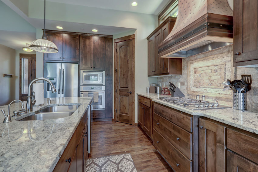 Kitchen with island, sink, faucet, countertops, range hood, cabinets, wood flooring, and stacked stone backsplash