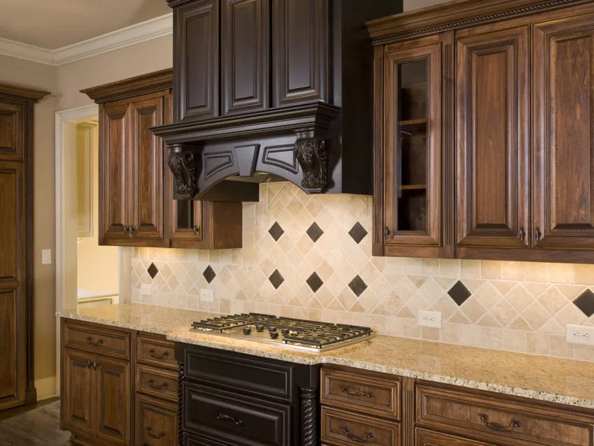 Kitchen with travertine backsplash, wood cabinets, range hood, countertop, and stove