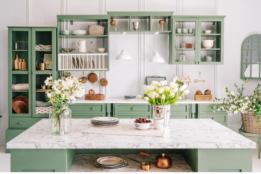 Kitchen with vintage design counter with marble top and flowers in metal bucket on it, and organized pastel cabinets with various crockery