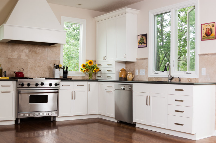 Kitchen with white cabinets, travertine backsplash, wood flooring, oven, countertop, dishwasher, and window