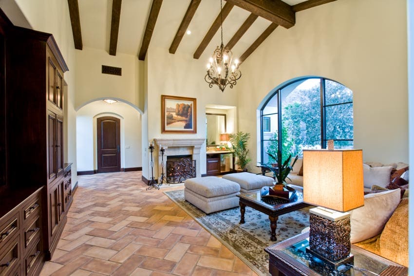 Living room with modern terracotta tile floor, lamp, ceiling beams, windows, chandelier, cabinets, and couch