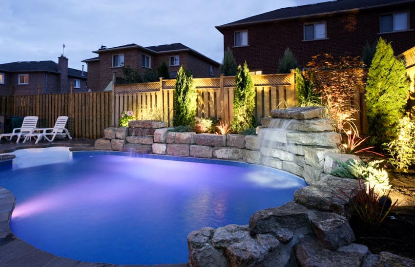 Outdoor pool area with water fixtures, lights, lounge chairs, and wooden fence