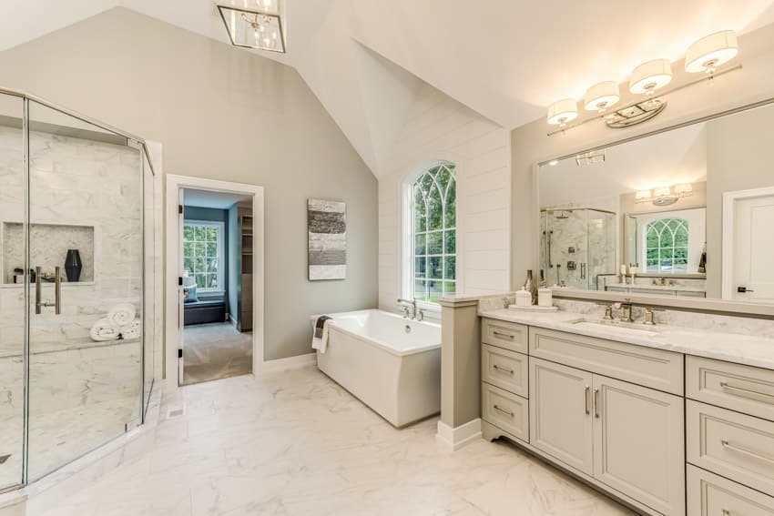 Spacious bathroom with glass door, shower, Carrara marble floors, walls, mirror, tub, window, and cabinets