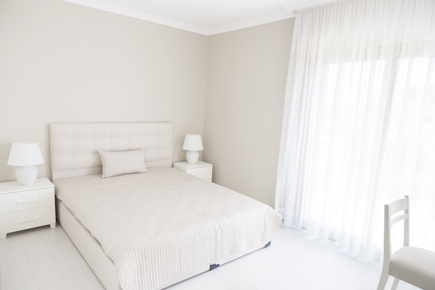 White bedroom with headboard, pillows, nightstands, lamps, chair, curtains, and flat sheet bedding